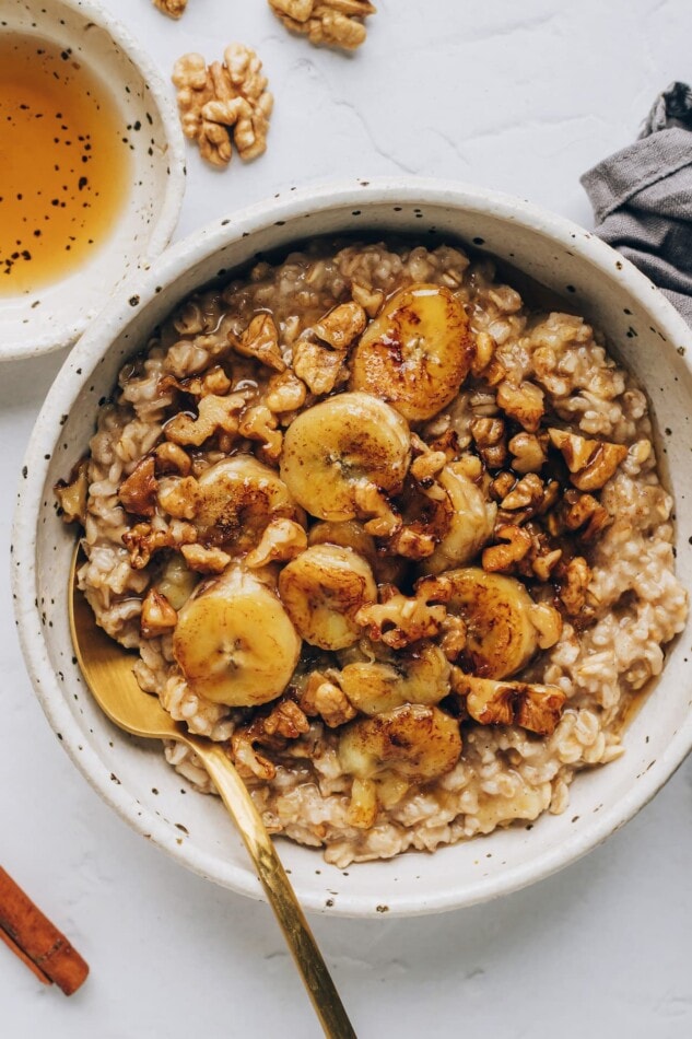 Banana bread oatmeal in a white bowl with caramelized bananas and toasted walnuts on top. Gold spoon and coffee mug in the background. An overhead photo of a bowl of banana bread oatmeal, topped with banana slices and walnuts, and drizzled with maple syrup.