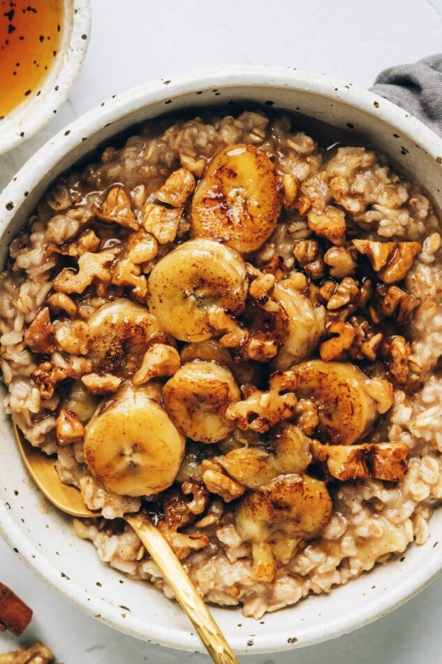 Banana bread oatmeal in a bowl placed on a natural napkin with a gold spoon. An overhead photo of a bowl of banana bread oatmeal, topped with banana slices and walnuts, and drizzled with maple syrup.