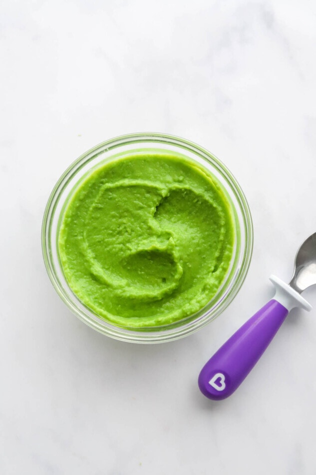 A small jar of pea puree. A purple spoon is cut out of the frame.