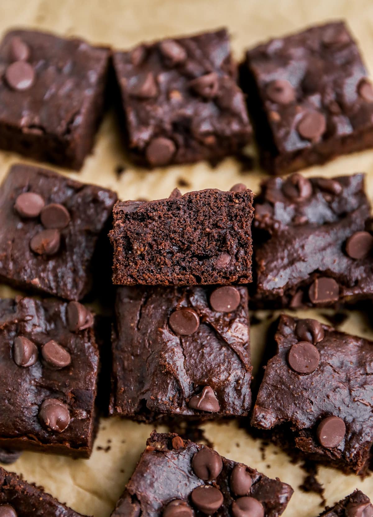 Squares of protein brownies on a sheet of brown parchment paper. The center brownie is resting against another brownie, exposing the inside.