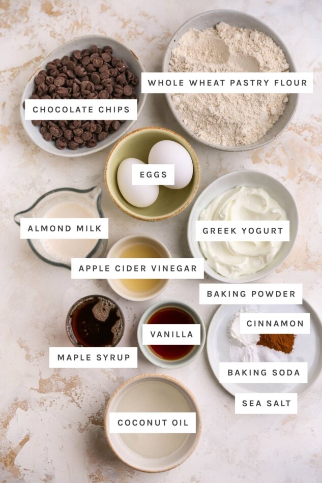 Ingredients measured out to make healthy chocolate chip muffins: chocolate chips, whole wheat pastry flour, eggs, Greek yogurt, almond milk, vinegar, baking powder, cinnamon, vanilla, baking soda, sea salt, maple syrup and coconut oil.