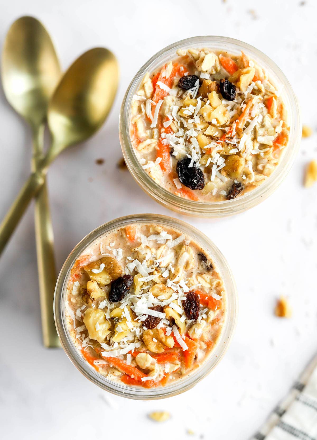 carrot-cake-overnight-oats-5.jpg An overhead view of two jars containing carrot cake overnight oats. Two gold spoons rest next to the jars.