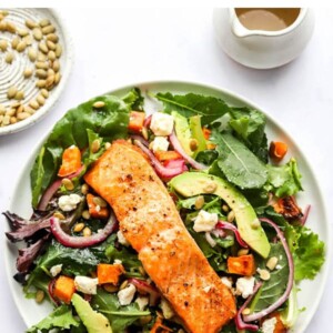 Salmon salad on a plate topped with salmon, red onions, avocado, sweet potatoes, pepitas and feta.