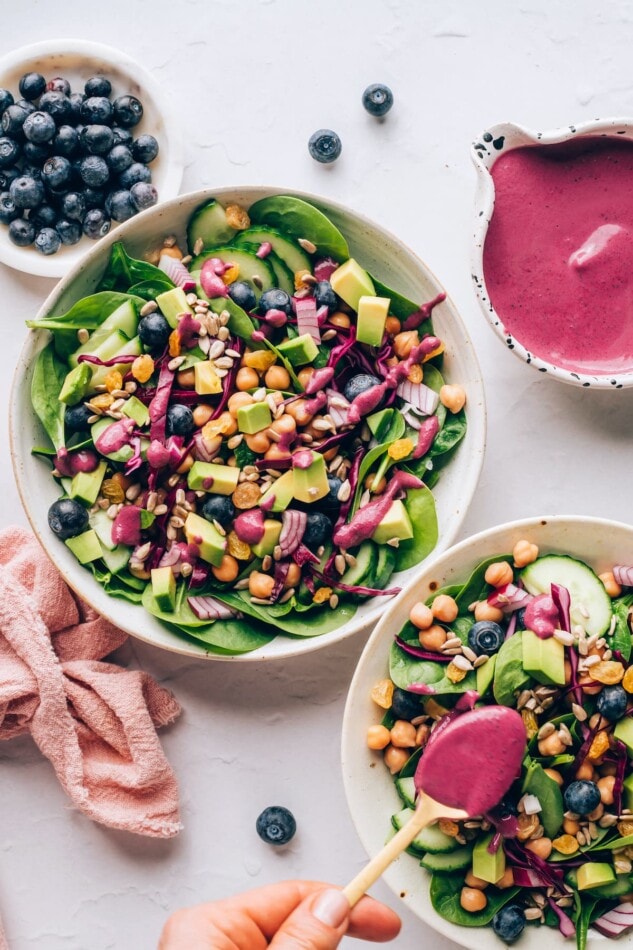 Antioxidant Salad with Blueberry Tahini Dressing #vegan #healthy #plantbased An overhead photo of two bowls of spinach blueberry salad. The salad in the bottom right corner is being drizzled with blueberry tahini dressing.