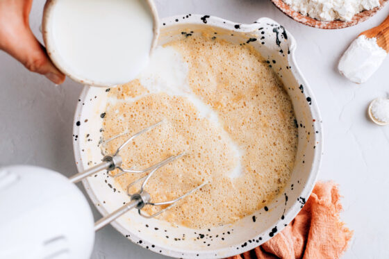 Almond milk and vinegar mixture added to a butter mixture in a large mixing bowl with a hand mixer in the bowl.