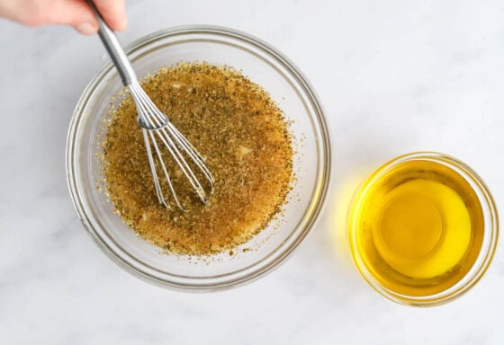 Vinegar, lemon juice, water, dijon mustard, honey, dried oregano, garlic, sea salt and black pepper whisked together in a bowl. A small bowl of olive oil is nearby.