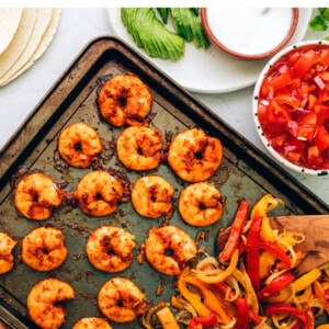 Cooked veggies and shrimp on a sheet pan. Fajitas accoutrements surround the sheet pan.