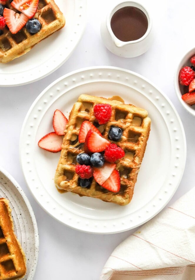 An almond flour waffles on a white plate. It has been topped with various berries and a drizzle of maple syrup.