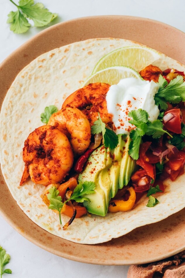 These sheet pan shrimp fajitas make for an easy weeknight meal with Mexican flavor. Simply bake your shrimp, peppers and onions together on one sheet, load up your tortillas and enjoy! A shrimp fajita on a plate topped with avocado slices and greek yogurt.
