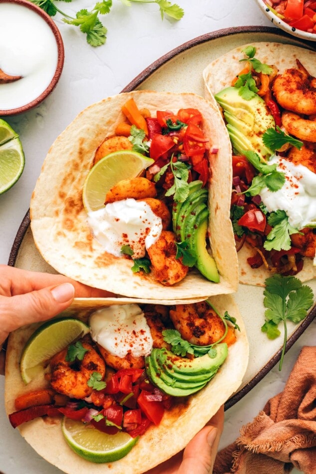 These sheet pan shrimp fajitas make for an easy weeknight meal with Mexican flavor. Simply bake your shrimp, peppers and onions together on one sheet, load up your tortillas and enjoy! Three fajitas on a serving plate. Hands are removing a fajita.