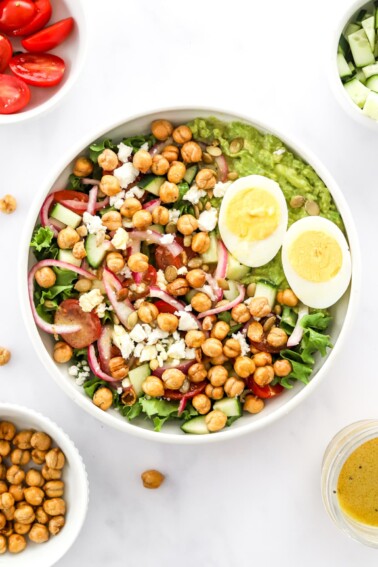 A large bowl of cooked chickpeas, hard boiled egg, lettuce, red onion, feta cheese, cherry tomatoes, and avocado.