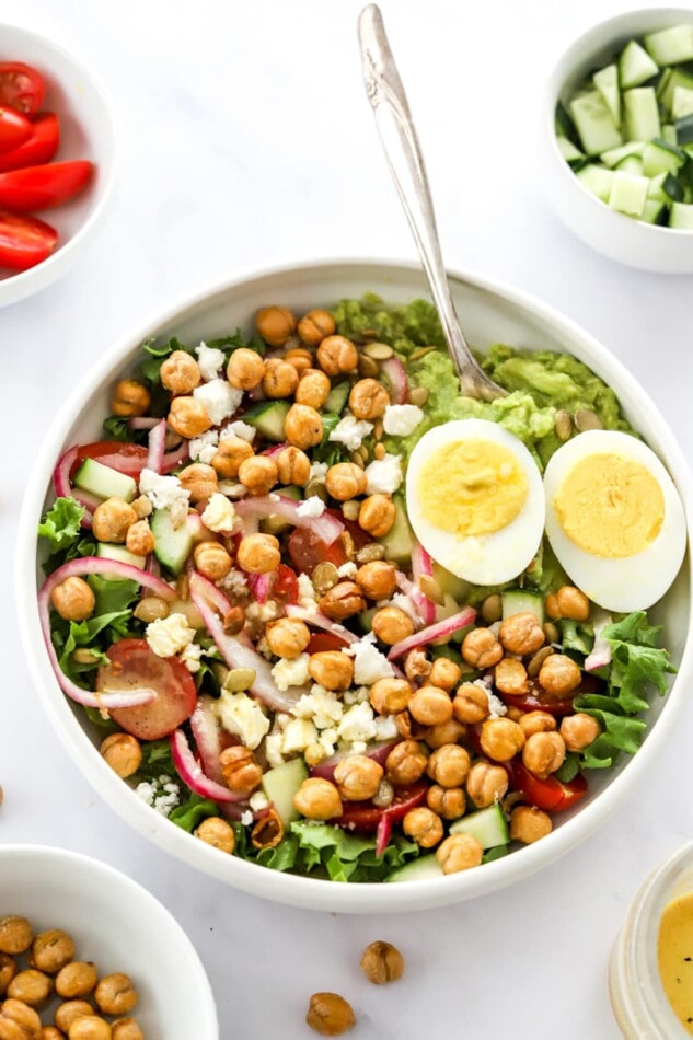 A serving of mashed avocado, a hard boiled egg, chickpeas, feta cheese, red onion, cucumber, spices, and cherry tomatoes.