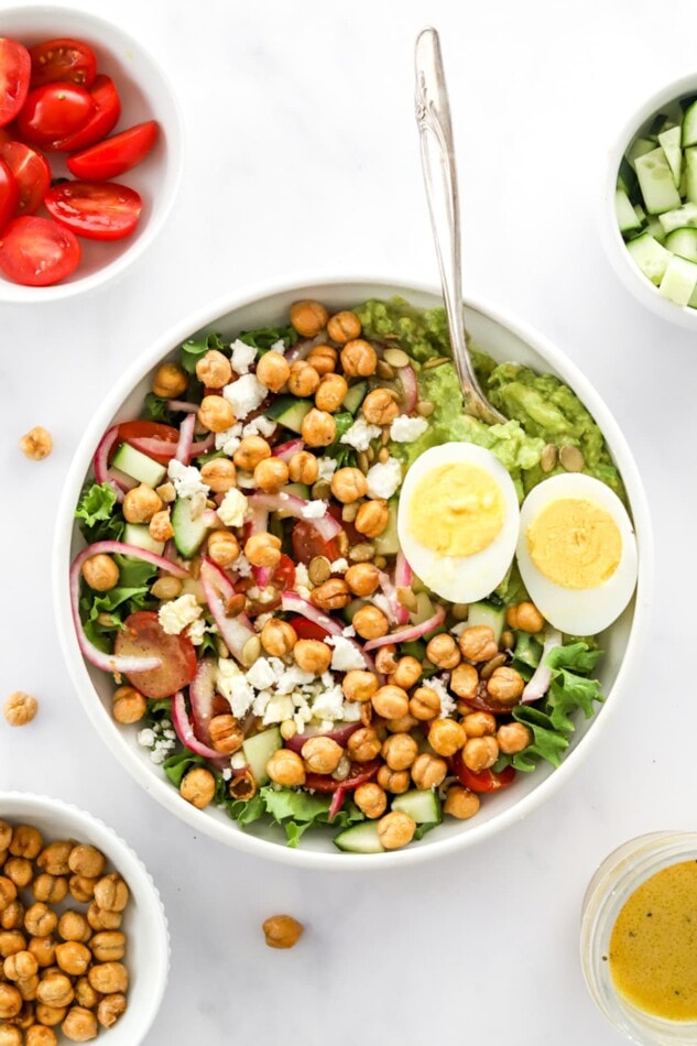 A serving of mashed avocado with a hard boiled egg, cooked chickpeas, feta cheese, red onion, lettuce, and cherry tomatoes in a bowl.