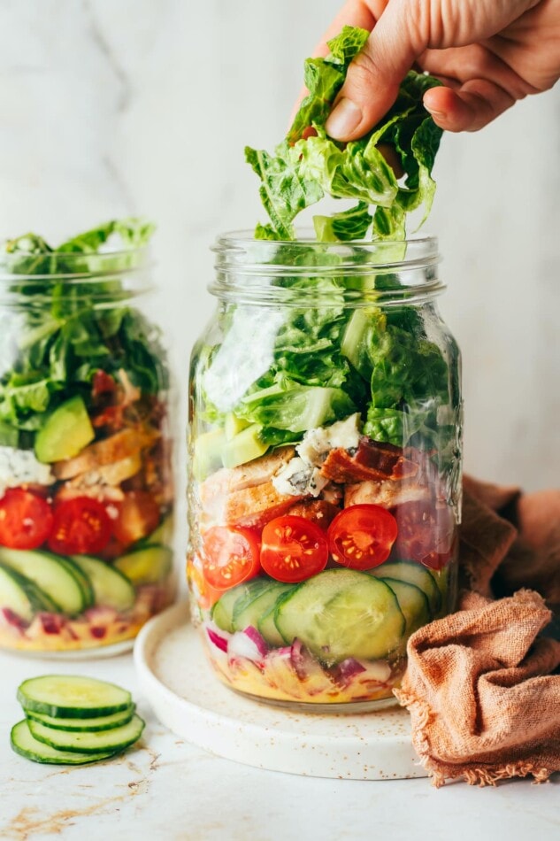 Two large mason jars with red onion, cucumber, cherry tomatoes, chicken, lettuce, feta cheese.