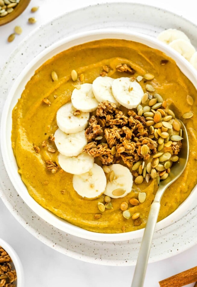 A pumpkin smoothie in a bowl with various toppings including banana slices, crunchy granola, and pumpkin seeds.