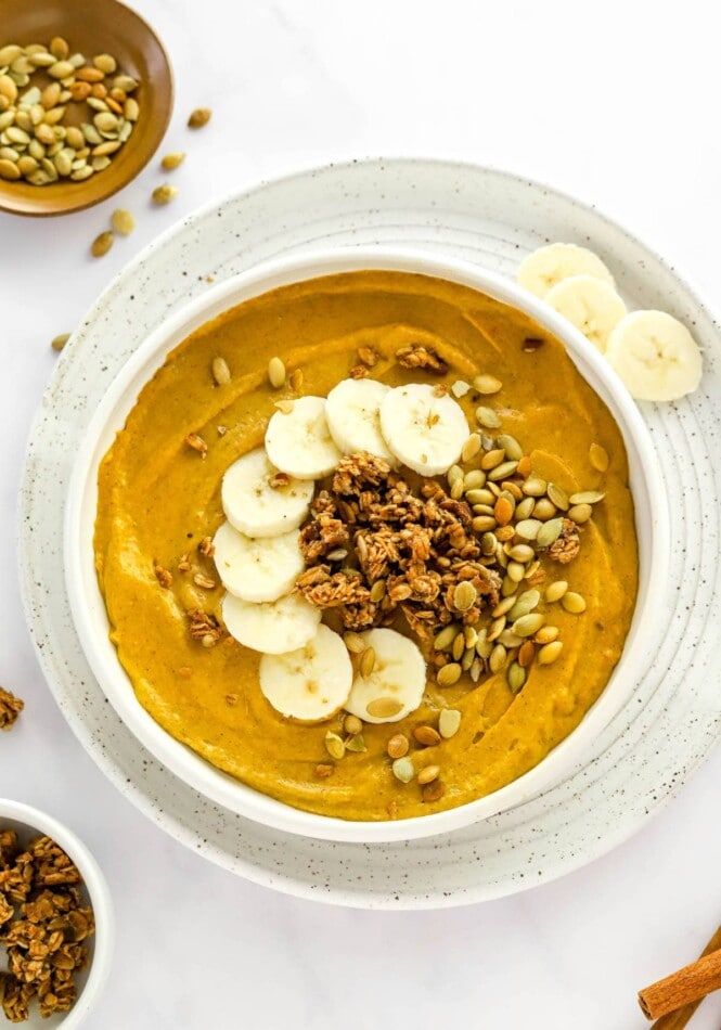 A serving of a pumpkin smoothie in a bowl, served on a plate, and topped with banana slices, pumpkin seeds, and granola.