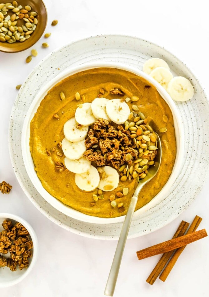 A pumpkin smoothie bowl served with banana slices, granola, and pumpkin seeds. The bowl is served on a plate with cinnamon sticks and a small bowl of granola off to the side.