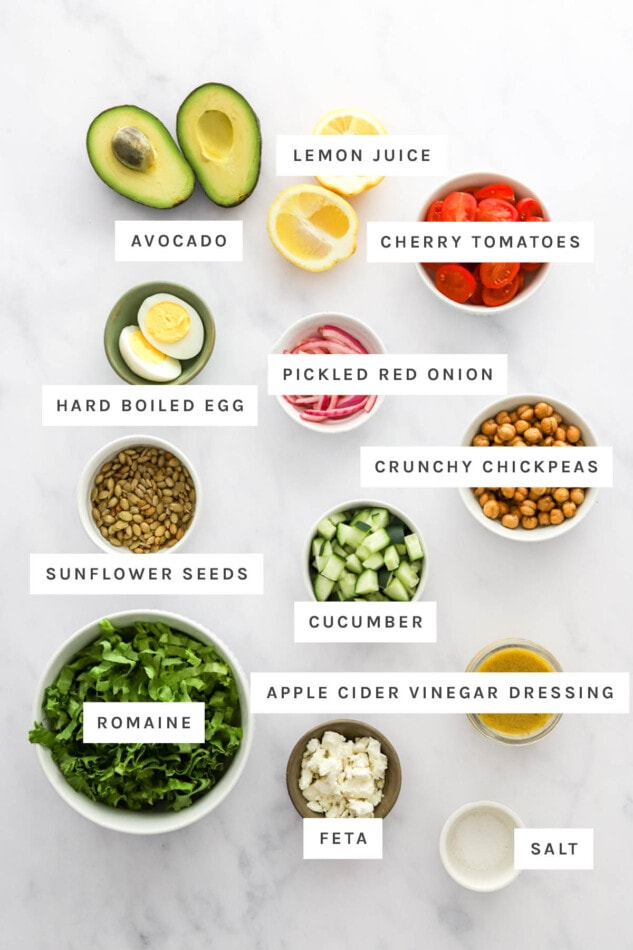 Ingredients measured out to make Mashed Avocado Bowl: avocado, lemon juice, cherry tomatoes, hard boiled egg, pickled red onion, crunchy chickpeas, sunflower seeds, cucumber, vinegar dressing, romaine, feta and salt.