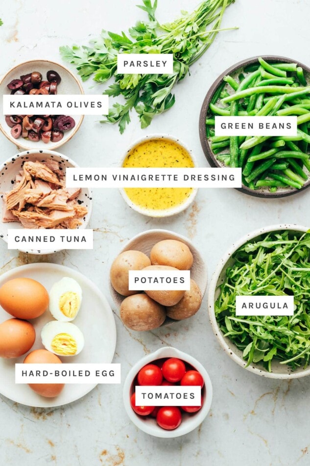 PIN THIS --> Meal Prep Nicoise Salad Ingredients measured out to make nicoise salad: parsley, kalamata olives, green beans, lemon vinaigrette dressing, canned tuna, potatoes, arugula, hard-boiled egg and tomatoes.