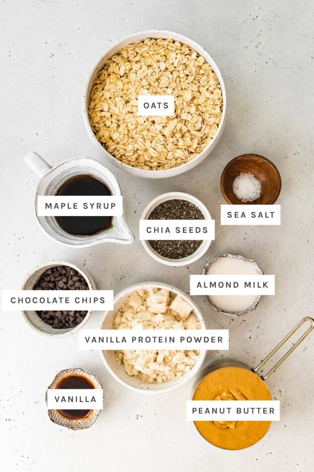 Ingredients measured out to make Peanut Butter Protein Bars: oats, maple syrup, sea salt, chia seeds, almond milk, chocolate chips, vanilla protein powder, almond milk, vanilla and peanut butter.