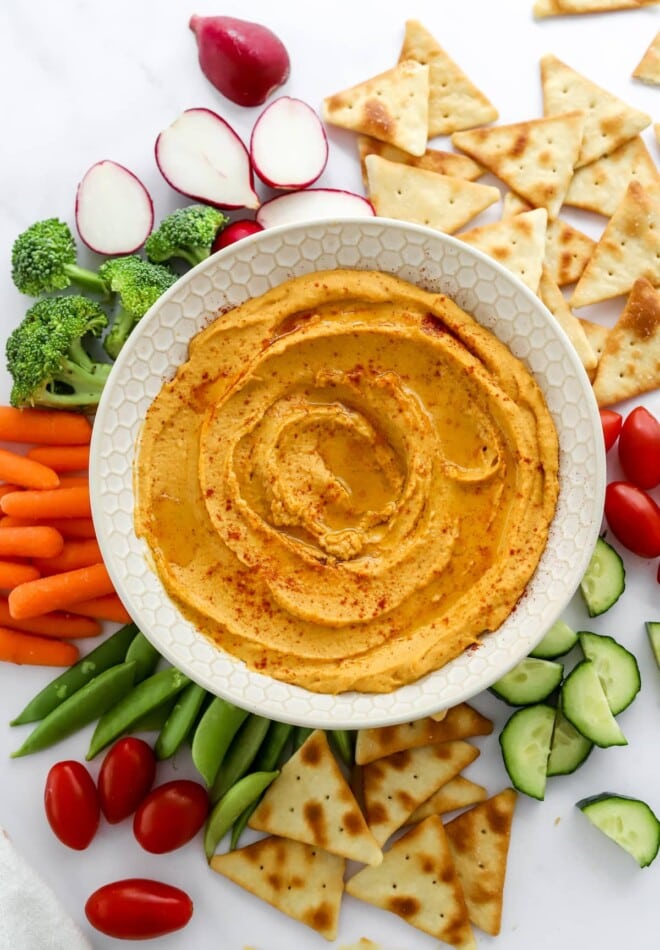 A bowl of sweet potato hummus surrounded by various vegetables and pita chips for dipping.