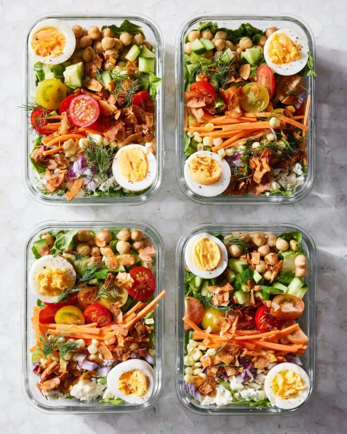 Four glass meal prep containers with salads that contain hard boiled eggs, beans, tomatoes, carrots, cucumbers, onion, lettuce, and fresh herbs.