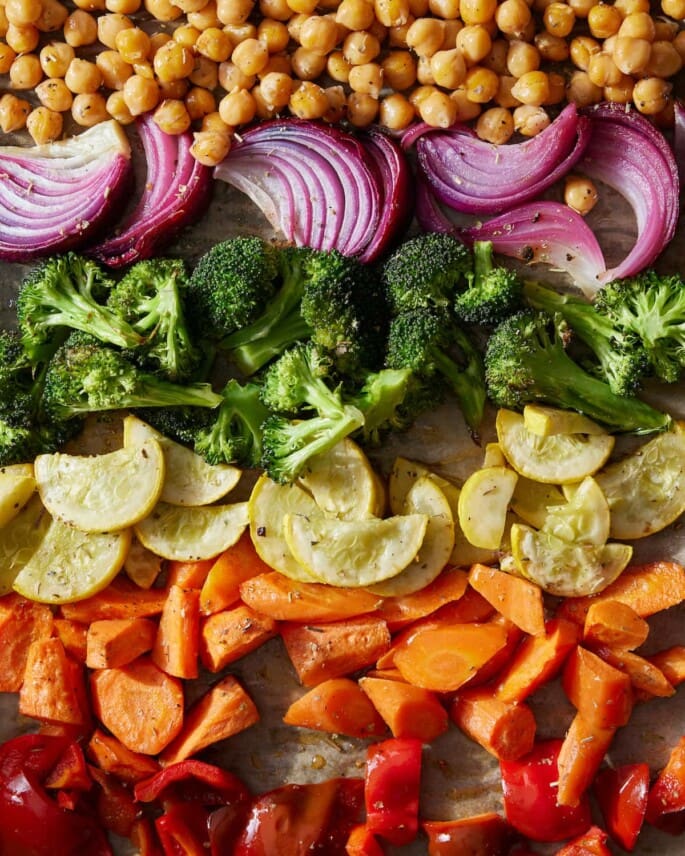 A sheet pan with rows of roasted chickpeas, red onion, broccoli, yellow squash, carrots, and red bell pepper.