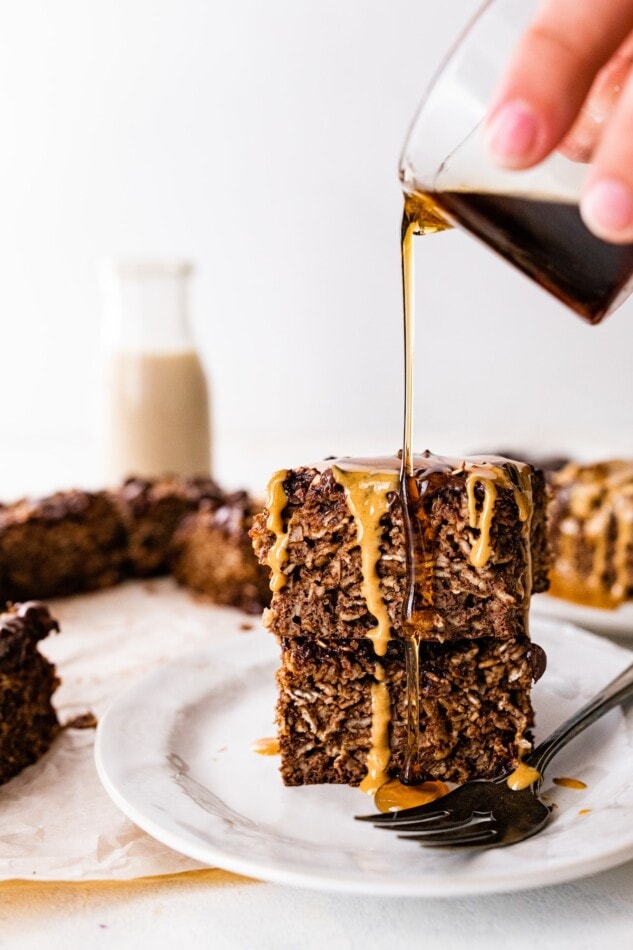 Two portions of baked oatmeal stacked on top of each other, a hand is pouring maple syrup out of a container onto the oatmeal.