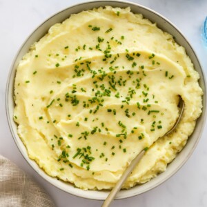 A large bowl of mashed potatoes garnished with fresh chives and a metal spoon in the mashed potatoes.