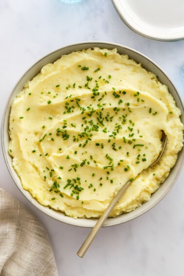 A large bowl of mashed potatoes garnished with fresh chives and a metal spoon in the mashed potatoes.
