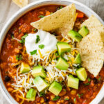 A bowl of taco soup topped with avocado chunks, shredded cheese and sour cream with tortilla chips.