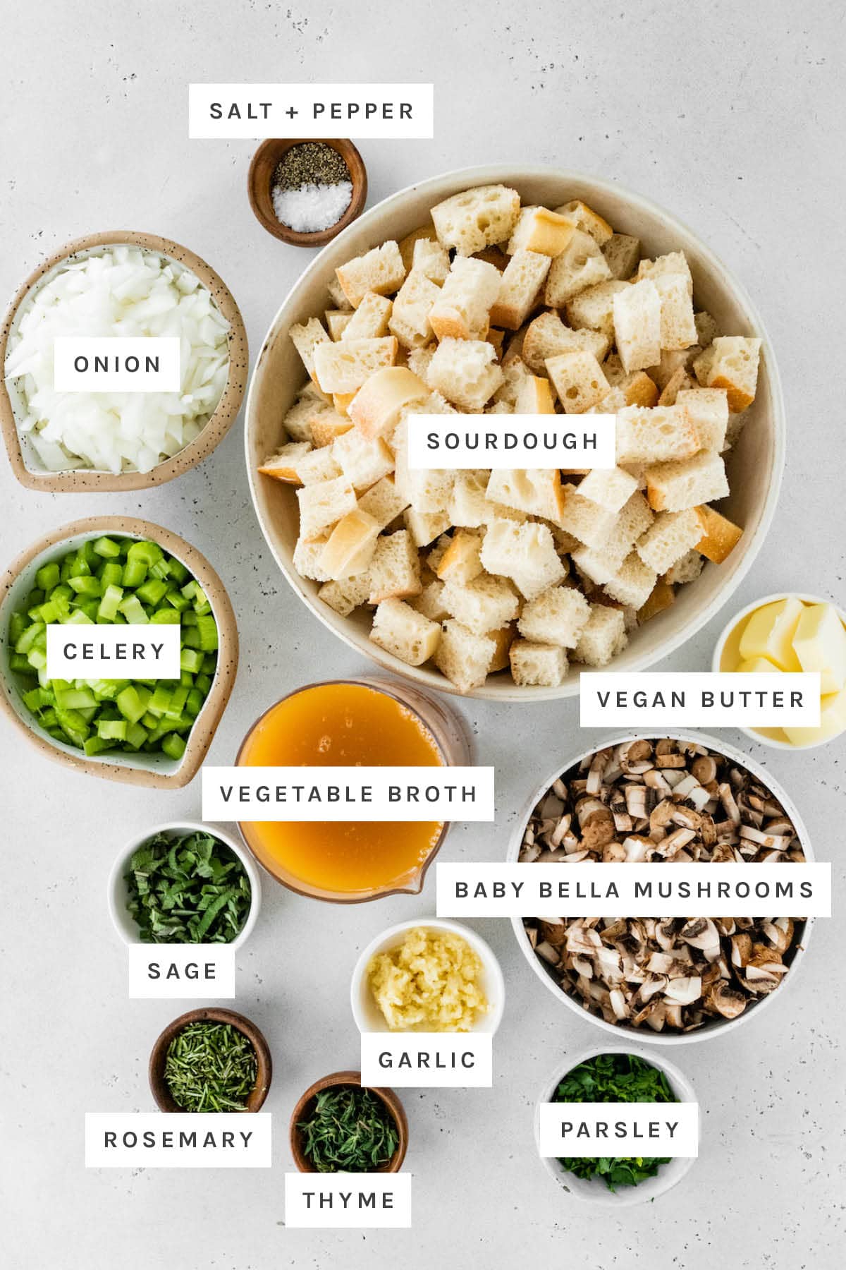 Ingredients measured out to make Sourdough Stuffing: salt, pepper, onion, sourdough, celery, vegetable broth, vegan butter, sage, baby bella mushrooms, garlic, rosemary, thyme and parsley.