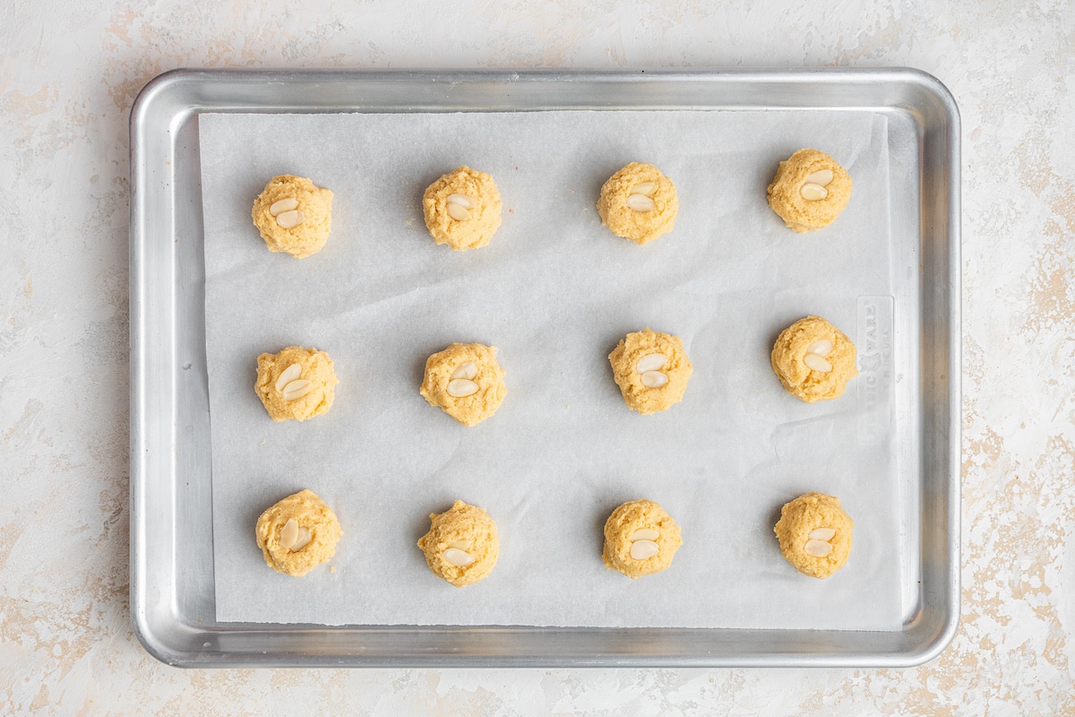 Almond cookie dough shaped into 12 balls on a parchment lined baking sheet topped with almond slivers.