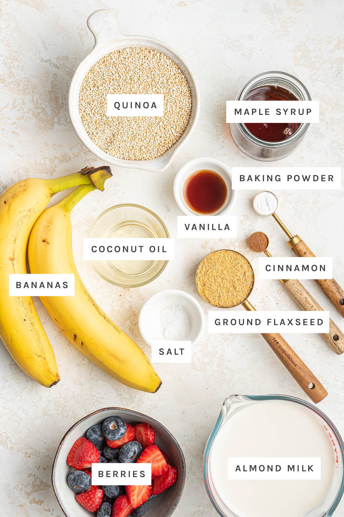Ingredients measured out to make Quinoa Breakfast Bake: quinoa, maple syrup, baking powder, vanilla, coconut oil, cinnamon, bananas, ground flaxseed, salt, berries and almond milk.