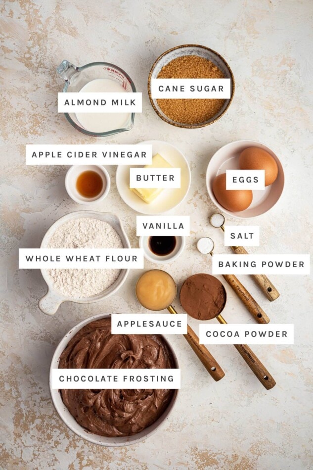Ingredients measured out to make Healthy Chocolate Cupcakes: almond milk, cane sugar, vinegar, butter, eggs, vanilla, whole wheat flour, salt, baking powder, applesauce, cocoa powder and chocolate frosting.