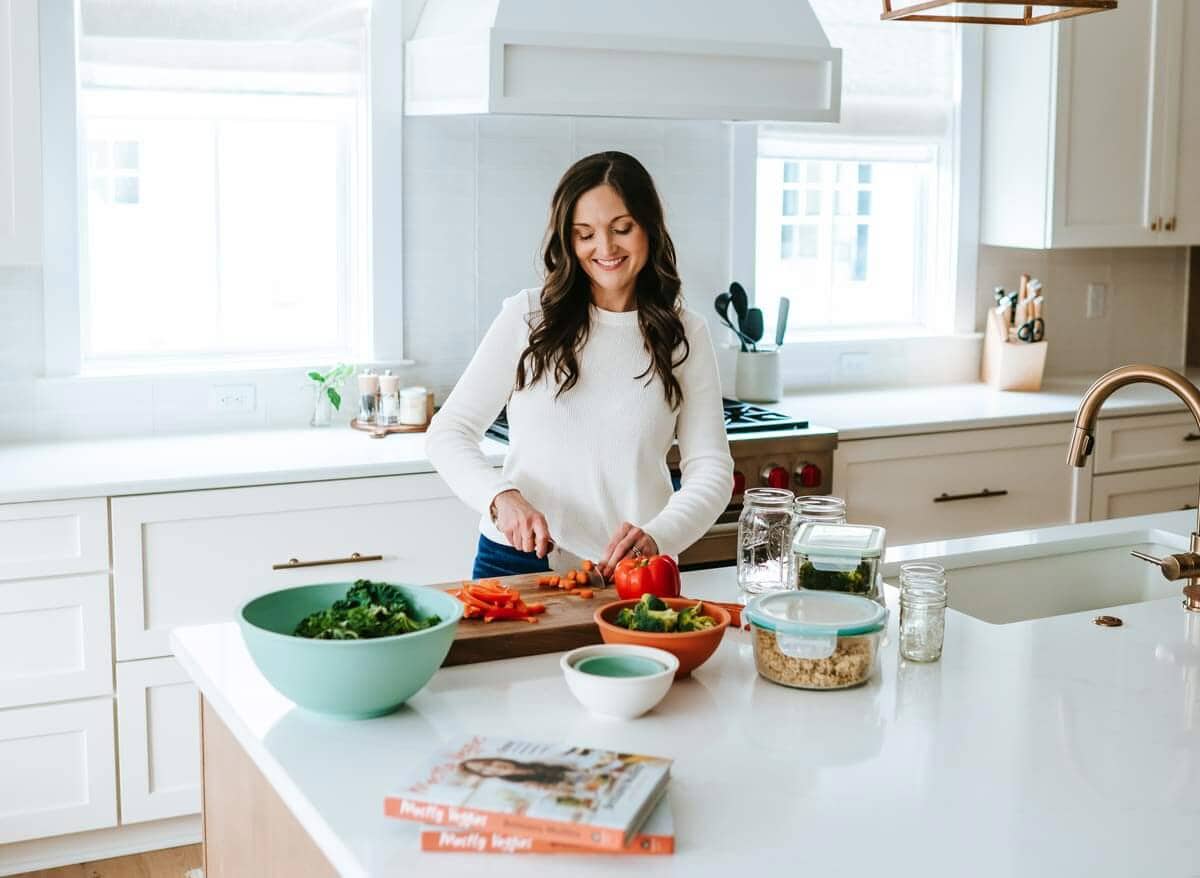 Brittany Mullins stands in her kitchen smiling while cutting vegetables on a wooden cutting board.