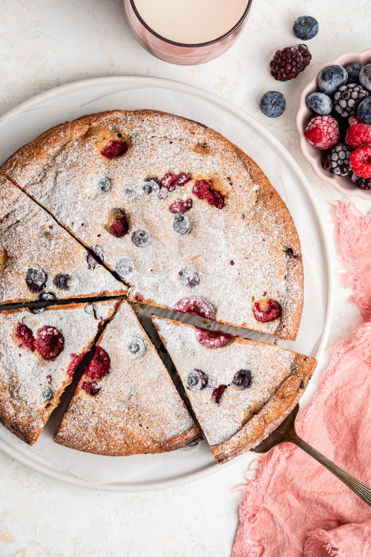 An easy berry cake cut into slices served on a white plate. A silver spatula is removing a slice of the cake from the plate.