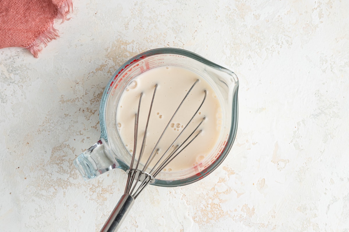 Almond milk, and vinegar mixed with a metal whisk in a glass measuring cup.
