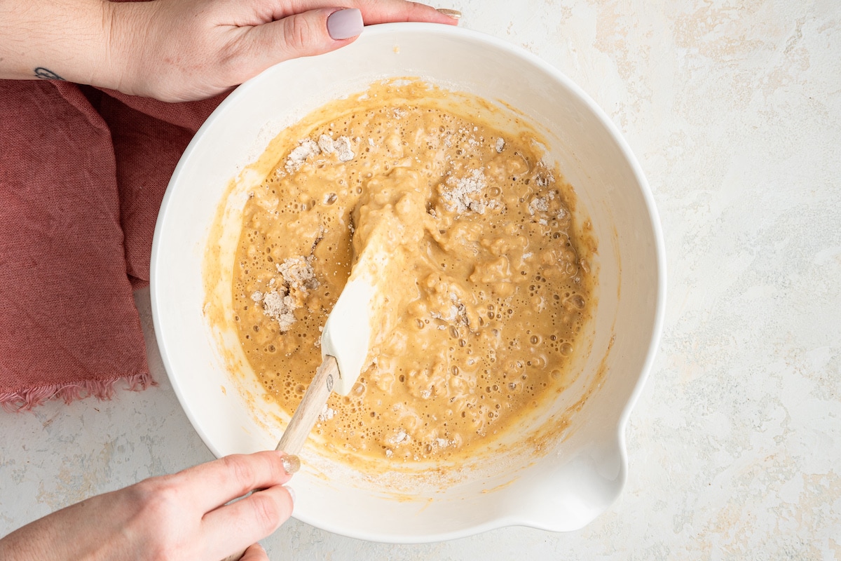 A woman's hand mixing the dry and wet ingredients together for the berry cake in a white mixing bowl.