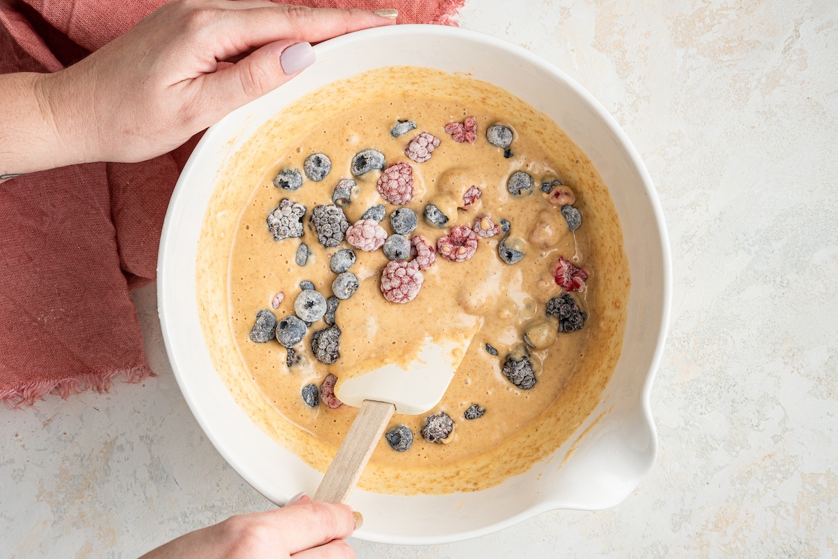 A woman's hand mixing berries into the berry cake batter with a spatula.