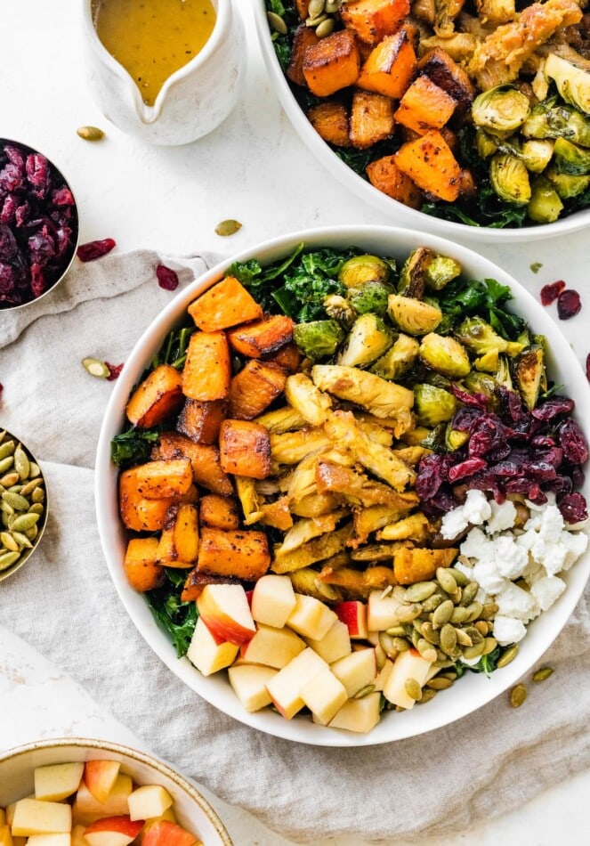 A serving of a roasted harvest bowl with sweet potato, Brussels sprouts, dried cranberries, apples, pumpkin seeds, feta cheese, and kale.