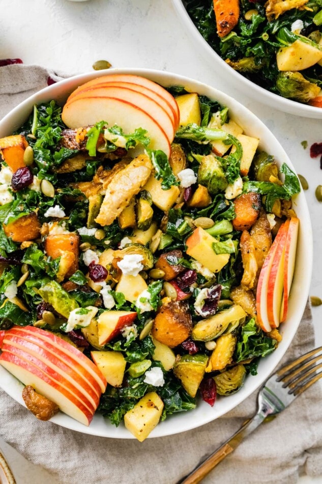 A bowl of a harvest salad with sweet potato, apples, feta cheese, Brussels sprouts, dried cranberries, and pumpkin seeds.
