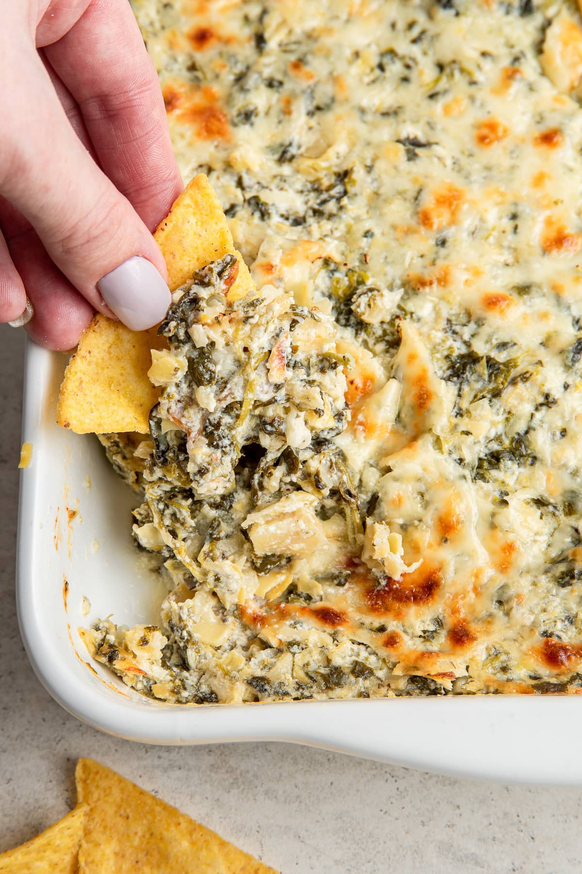 A hand dipping a chip into a dish of spinach artichoke dip.