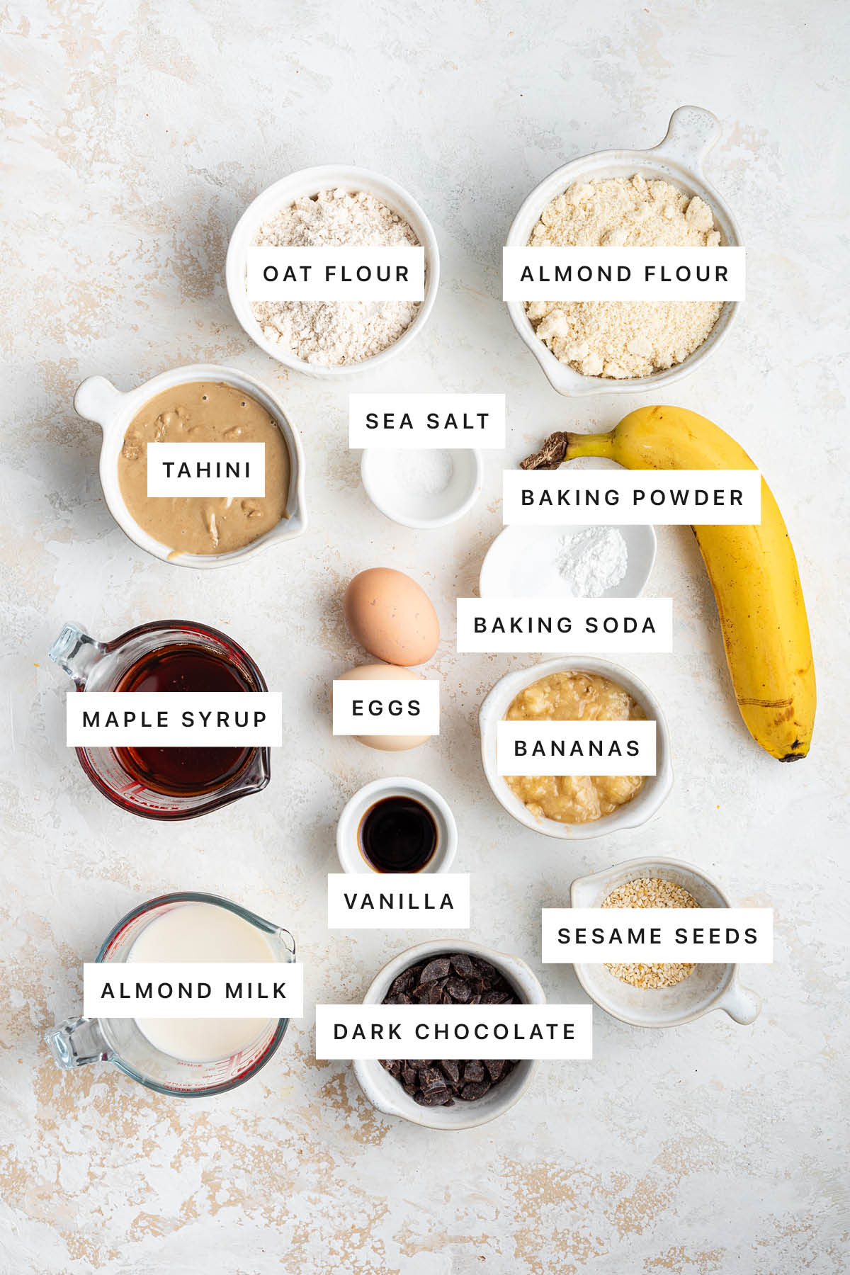 Ingredients measured out to make Tahini Banana Bread: oat flour, almond flour, sea salt, tahini, baking powder, baking soda, maple syrup, eggs, bananas, vanilla, almond milk, dark chocolate and sesame seeds.