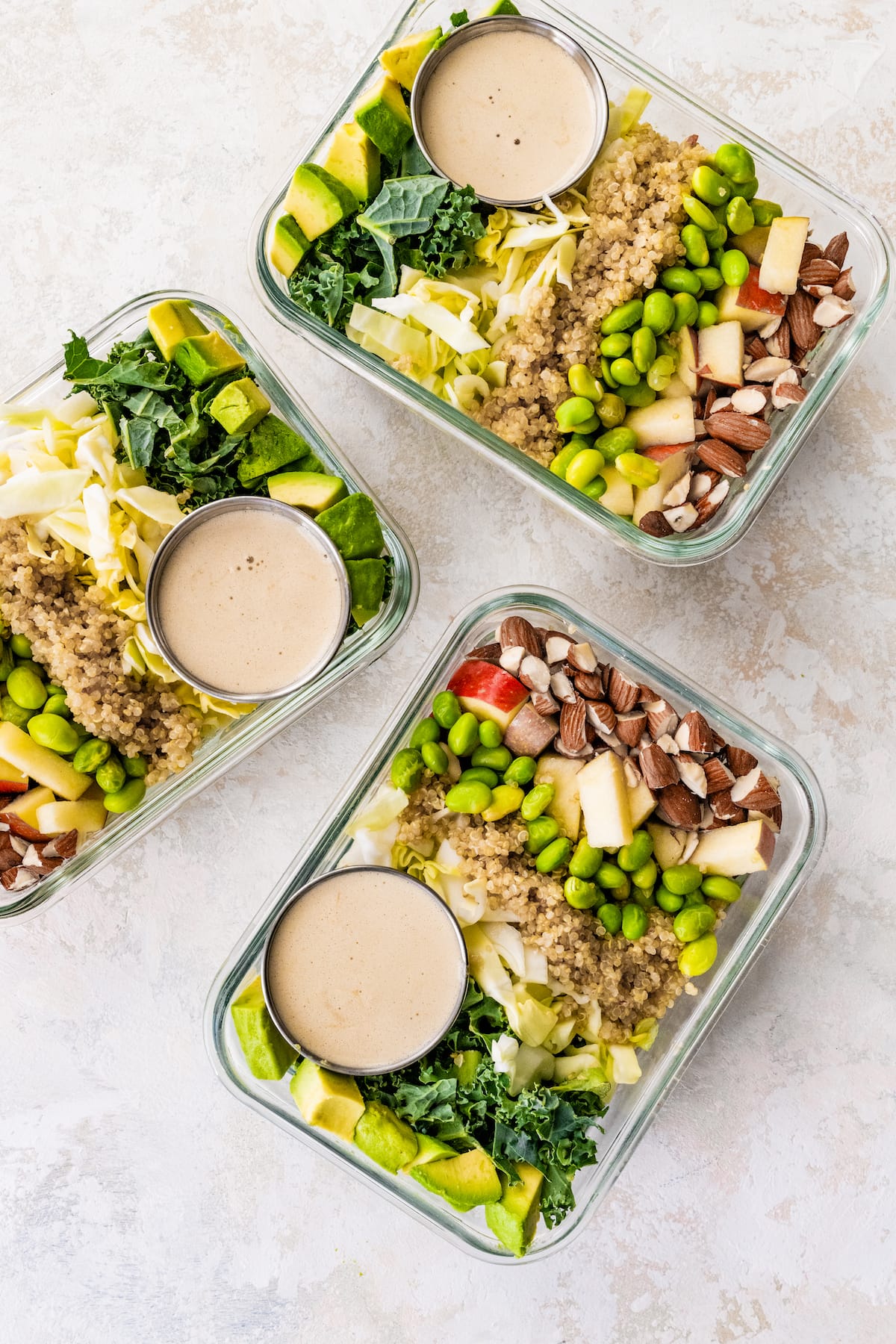 Three glass storage containers layered with detox salad ingredients: almonds, apple, edamame, quinoa, cabbage, kale and avocado. A small container of dressing is also in each of the containers.