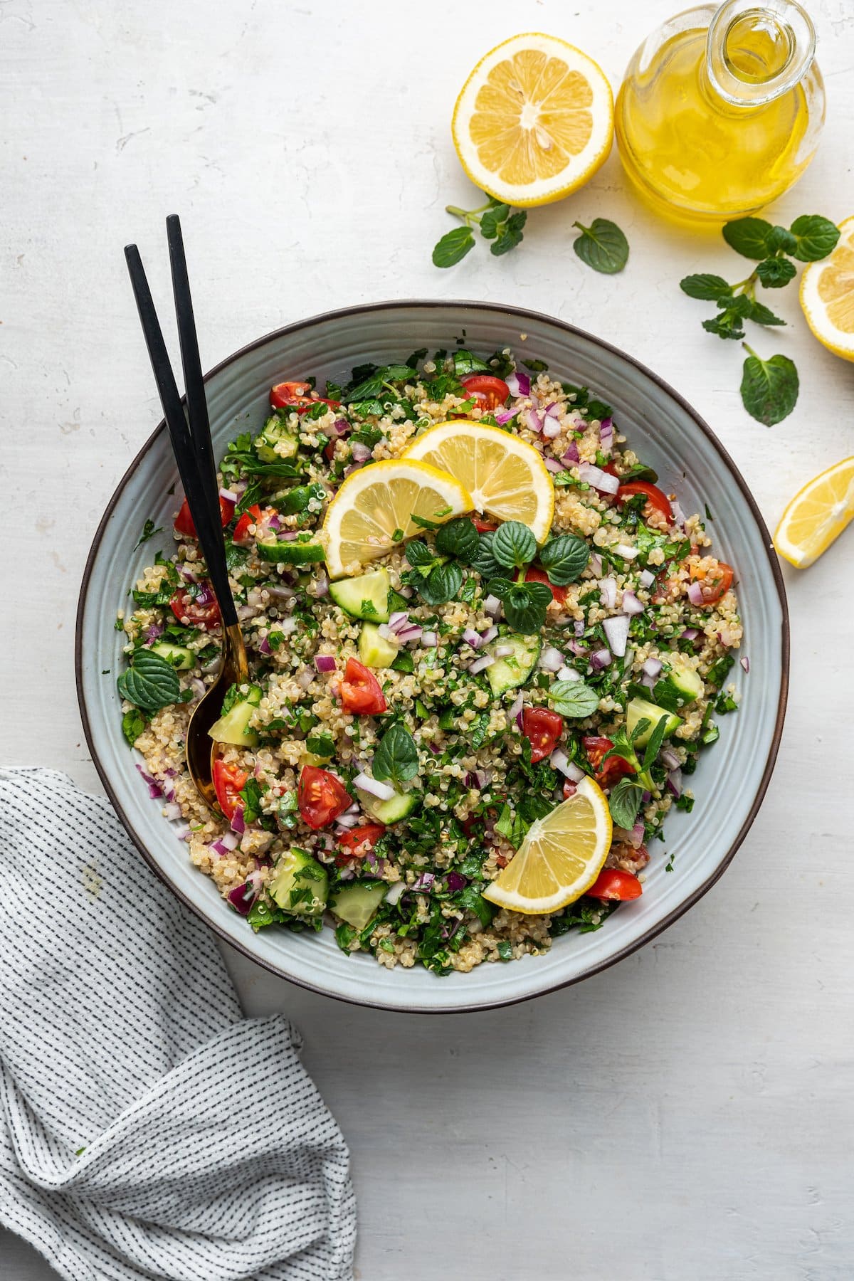 PIN THIS--> Quinoa Tabbouleh Quinoa tabbouleh in a serving bowl with serving spoons topped with fresh lemon slices and fresh herbs.