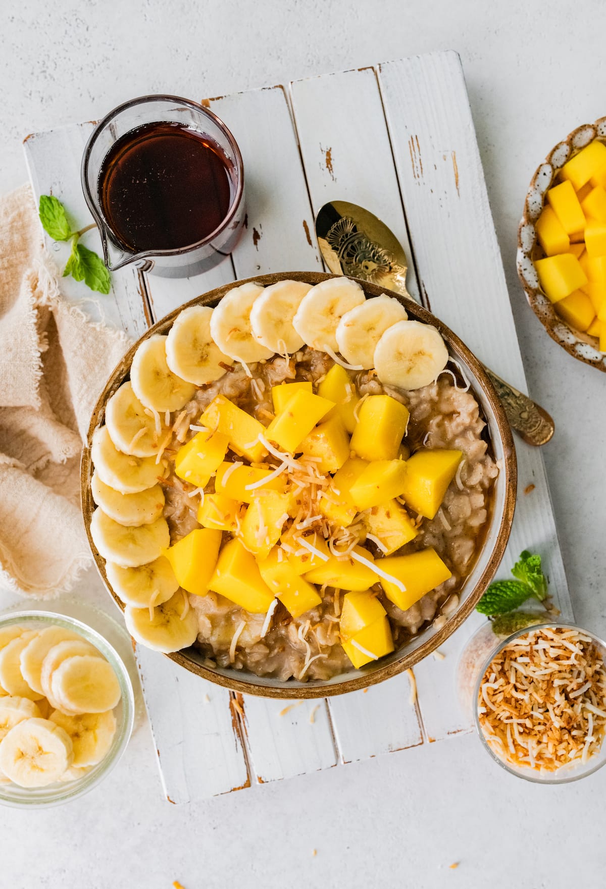 Tropical oats with banana, coconut and a drizzle of maple syrup. Far away bowl of oatmeal topped with fresh mango, banana slices and toasted coconut.