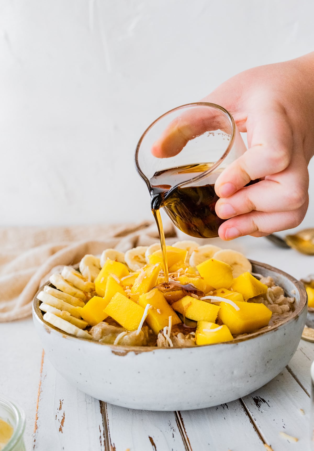 Tropical oats with banana, coconut and a drizzle of maple syrup. Woman's hand pouring maple syrup over bowl of tropical oatmeal.