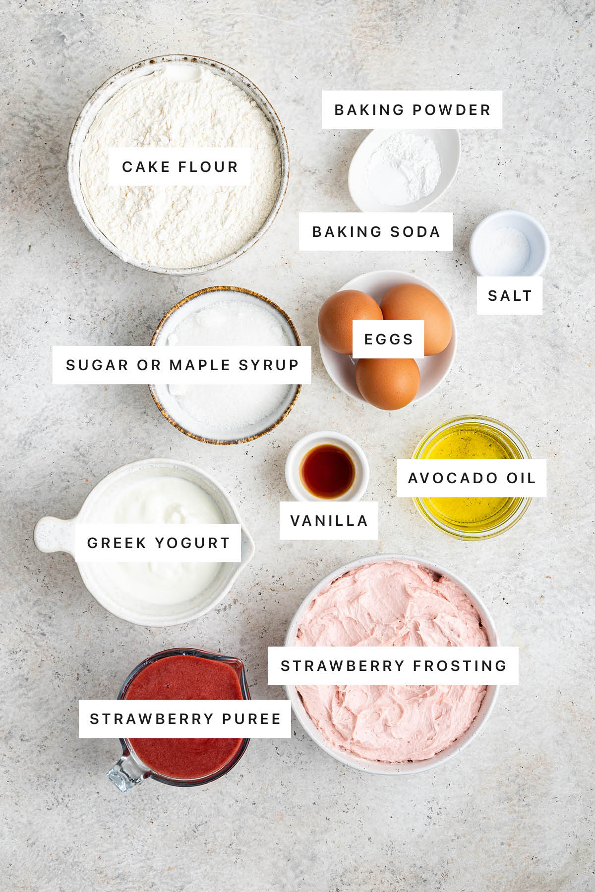 Ingredients measured out to make Healthy Strawberry Cake: cake flour, baking powder, baking soda, salt, eggs, sugar, vanilla, avocado oil, Greek yogurt, strawberry puree and strawberry frosting.
