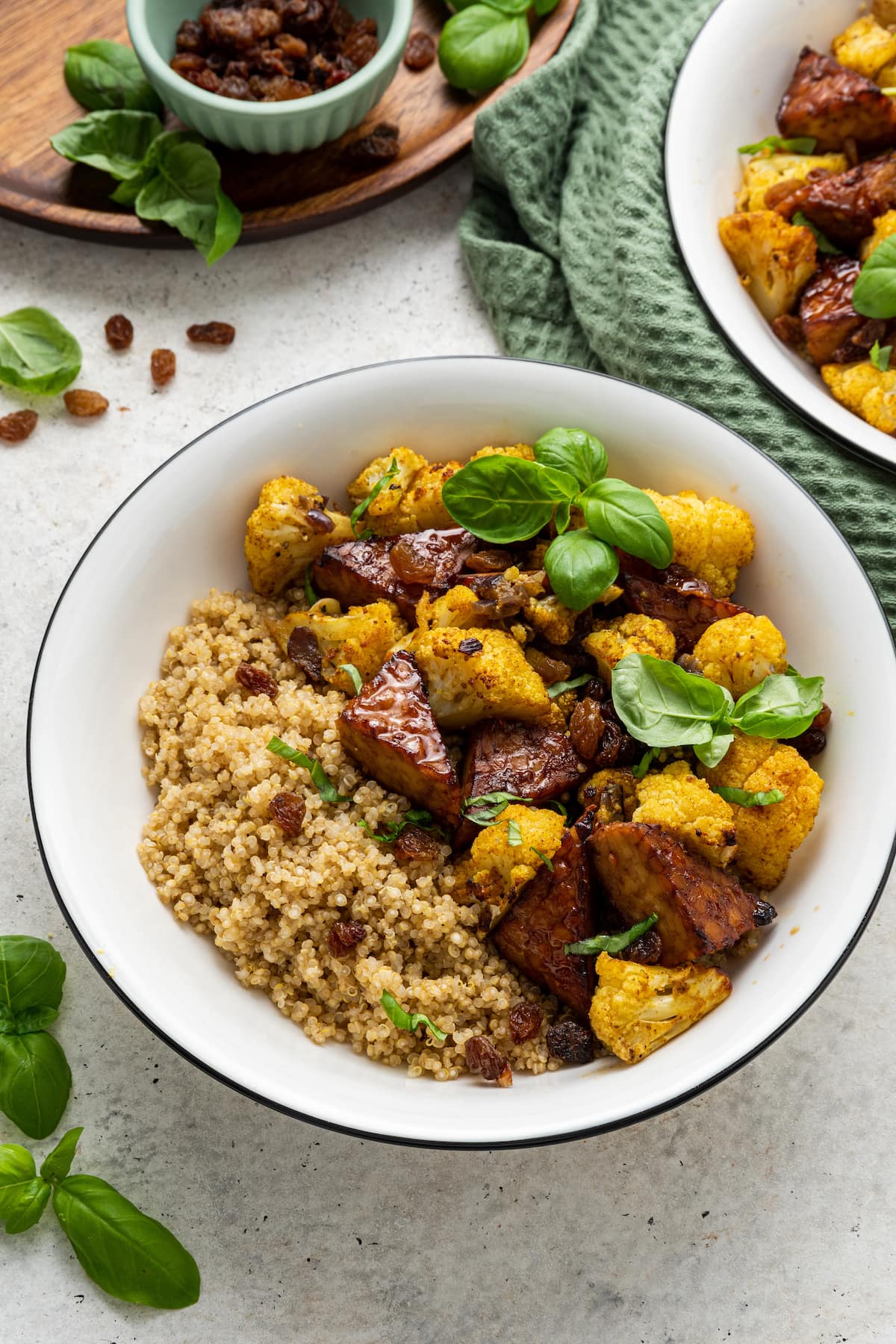 A white bowl containing the curried cauliflower and tempeh over a bed of quinoa and garnished with fresh basil leaves.